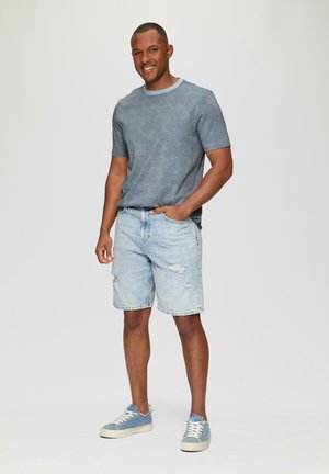 Hellblaues T-Shirt mit Rundhalsausschnitt aus strukturiertem Stoff, kombiniert mit hellblauen, ausgewaschenen Jeansshorts und blauen Sneakers mit weißen Sohlen.