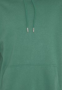 Détail avant d'un sweat à capuche vert montrant les cordons avec embouts métalliques et la couture de la poche kangourou sur un tissu doux.