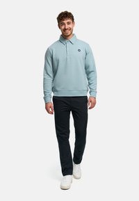 Lyseblå sweatshirt med polokrave og tre knapper, med et logo på venstre bryst, kombineret med mørke bukser og hvide sneakers.