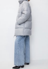 Chaqueta tipo puffer de color gris claro con un cuello alto, bolsillos laterales, forma cuadrada y textura acolchada, combinada con jeans de mezclilla desgastados y de pierna ancha, y zapatos negros.