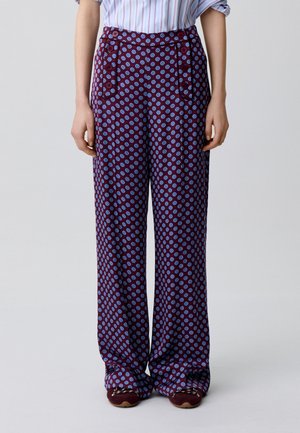 Donna che indossa pantaloni ampi color bordeaux con motivi circolari blu, abbinati a scarpe scure e una camicia a righe con maniche arrotolate.