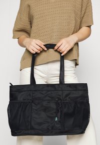 Bolsa tote negra de nailon con bolsillos laterales de malla dobles, cierre ajustable con cordón y un diseño elegante. Tejido resistente y asas reforzadas.