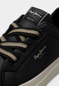 Baskets Pepe Jeans noires avec des lacets beige et noir rayés, semelle blanche et détails du logo sur la languette et le côté.