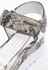 Sandal Calvin Klein s šedými hadími texturovanými popruhy a bílou polstrovanou stélkou na silné bílé podrážce.
