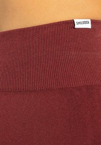 Bordeaux geribbelde tailleband met een gladde stof eronder, met een label "SMILODOX." Over het geheel een slank ontwerp, dat de textuur en kleur benadrukt.