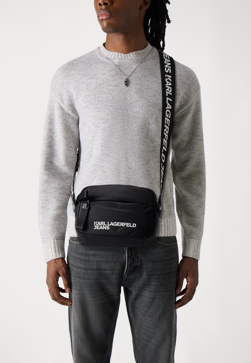 Schwarze Umhängetasche mit glatter Oberfläche, ausgestattet mit einem weißen Logo "KARL LAGERFELD JEANS" und einem verstellbaren Tragegurt mit Text.