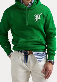 Grön hoodie med framficka och silverlogo, kombinerad med vita byxor och ett flerfärgat randigt bälte. Texturerat tyg, avslappnad passform.