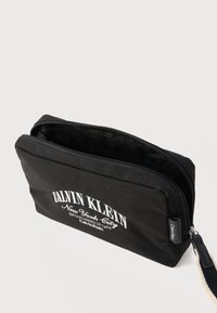 Μαύρο τσεπ άκι με φερμουάρ, λευκό λογότυπο Calvin Klein New York City και υφασμάτινο λουράκι καρπού σε λευκό φόντο.