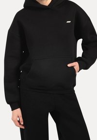 Personne portant un sweat à capuche noir et un pantalon noir, mains dans la poche du sweat, affichant une tenue décontractée avec un petit logo sur la poitrine.