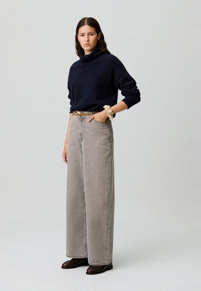 Suéter de cuello alto en marino, jeans de pierna ancha grises y zapatos marrones. El suéter tiene puños acanalados; los jeans tienen bolsillos frontales. Lleva una gomita en la muñeca.