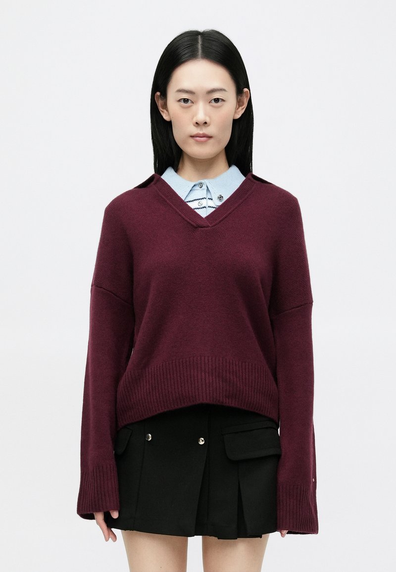 Burgundi színű kötött pulóver V nyakkal, széles ujjakkal és bordás szegéllyel, egy világoskék, csíkos mintás galléros ing fölé rétegezve.