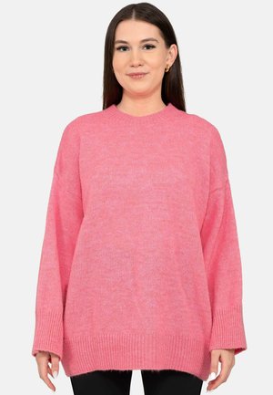 Young woman with straight brown hair wears oversized pink knit sweater and gold hoop earrings against plain light background.