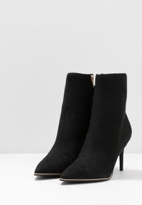 Paire de bottines en daim noir avec bouts pointus, talons hauts fins et fermetures éclair latérales, présentées sur une surface blanche.