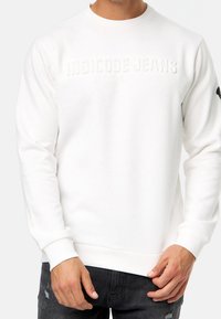 Hvid sweatshirt med præget "INDICODE JEANS" tekst, ribbede manchetter og kant, lavet af blødt bomuldsstof. Bæres med slidte mørke jeans.