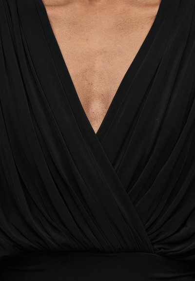 Zbliżenie osoby noszącej czarny strój z dekoltem w kształcie litery V, ozdobiony plisowanymi detalami z tkaniny na klatce piersiowej i ramionach.