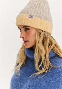 Dzianinowa czapka zimowa w jasnoszarym i żółtym kolorze, z prążkowaną fakturą, podwójnym mankietem i małą fioletową metką. Miękki materiał z mieszanki wełny.