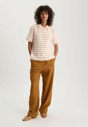 Femme debout, mains dans les poches, portant une chemise blanche à manches courtes avec des rayures rouges et un pantalon marron ample sur fond uni.