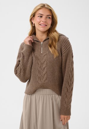 Jeune femme aux cheveux blonds portant un pull torsadé marron avec une fermeture éclair et une jupe beige, souriante et regardant sur le côté.
