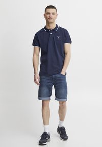 Mornarsko polo majico z belimi robovi, logotip na prsih. V kombinaciji s kratkimi jeans hlačami in temnimi športnimi čevlji. Model stoji z rokami v žepih.