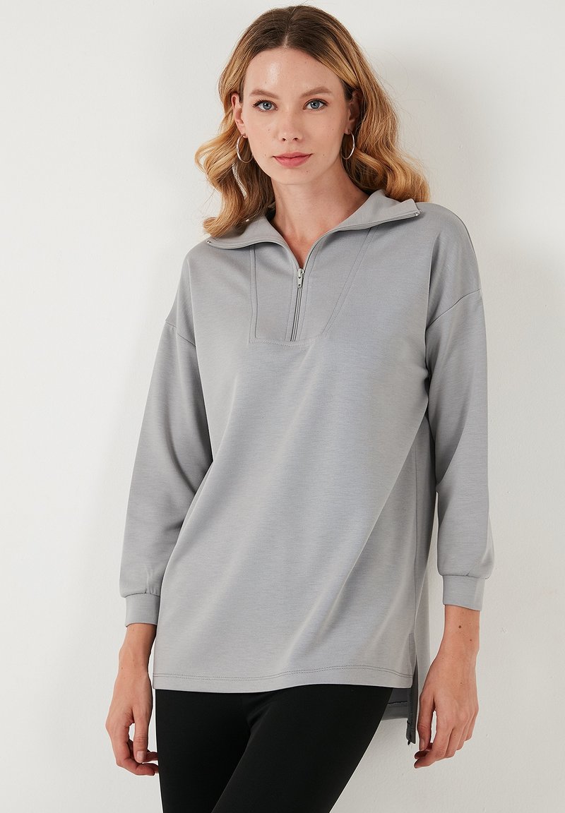Svetlo sivi pulover s half-zip ovratnikom, dolgimi rokavi in stranskimi razpoki. Gladka tekstura in sproščen kroj.