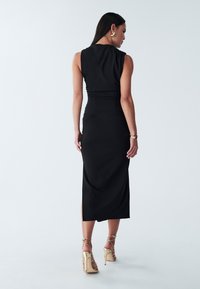 Abito midi nero con maniche a sbuffo, dettagli arricciati in vita e una texture liscia, abbinato a sandali alti con cinturini dorati.