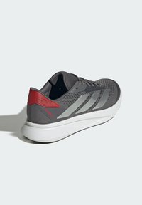 Zapatilla deportiva gris con parte superior de malla, acento rojo en el talón, franjas plateadas y suela blanca. Cuenta con cierre de cordones y talón reforzado para mayor soporte.