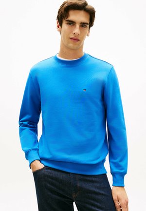 Jeune homme portant un sweat-shirt à col rond bleu vif et un jean foncé, debout avec une main dans la poche, sur un fond blanc uni.