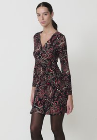 Vestido negro con un diseño de envolver, que presenta intrincados patrones en paisley rojo y dorado. Mangas largas y una forma ajustada, combinado con medias negras.