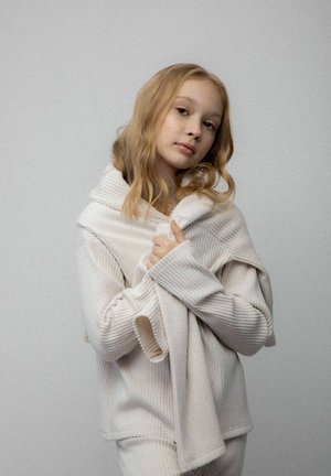 Młoda kobieta z blond włosami w kremowym, prążkowanym swetrze i spodniach, trzymająca kołnierz swetra obiema rękami na jednolitym szarym tle.