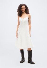 Νεαρή γυναίκα με σγουρά μαλλιά, ντυμένη με άσπρο midi φόρεμα χωρίς μανίκια και μαύρα μπότες μέχρι το γόνατο, να στέκεται σε απλό φωτεινό φόντο.