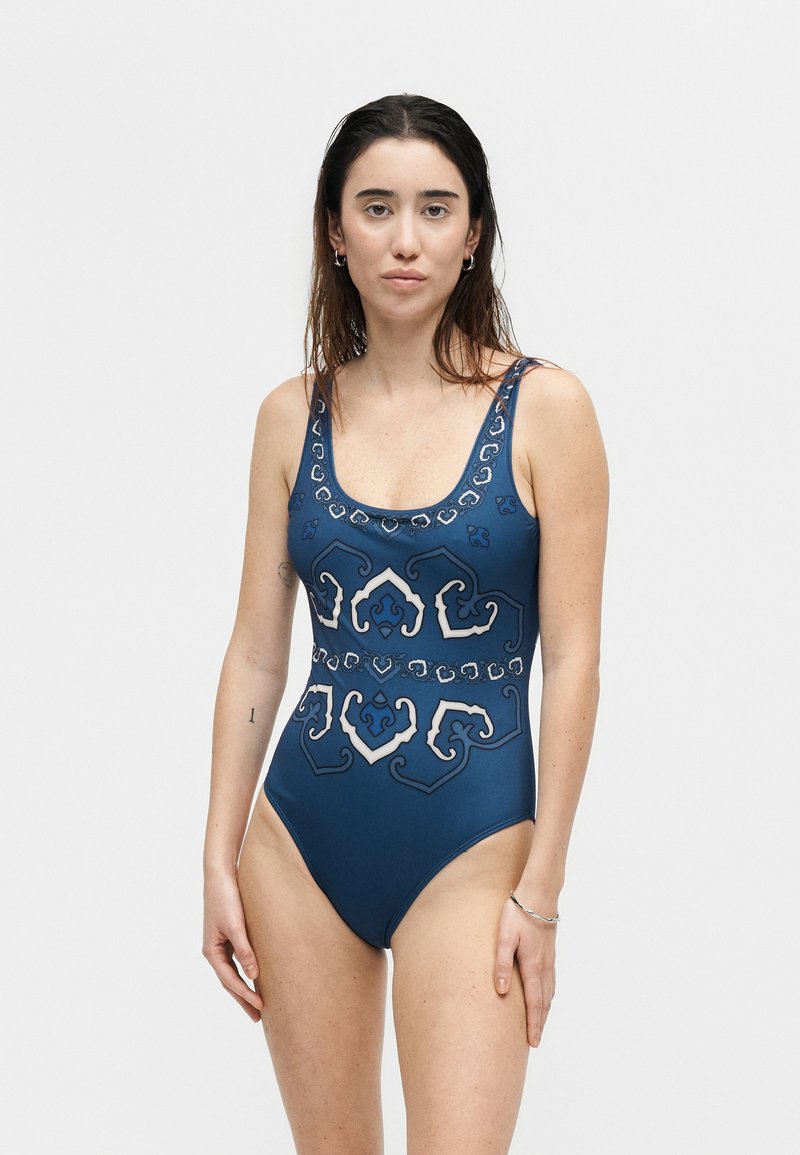 Woman wearing a blue one-piece swimsuit with white and black ornate patterns, standing against a plain light gray background.