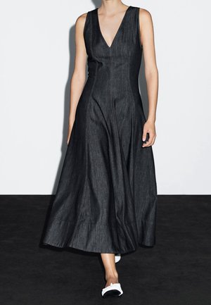 Woman wearing sleeveless, deep V-neck black denim maxi dress and white low-heeled shoes, standing against a plain white and black background.