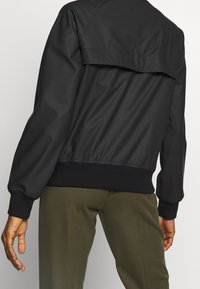 Osoba v černé bundě s žebrovanými manžetami a pasem, a olivově zelených kalhotách, zobrazená zezadu na jednobarevném pozadí.