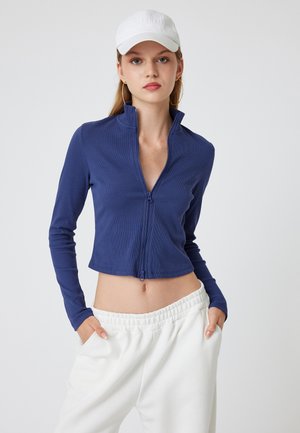Kobenblaues, tailliertes Zip-Up-Jacket mit langen Ärmeln und gerippter Textur, kombiniert mit weißen Sweatpants in lässigem Schnitt.