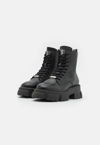 Botas de tobillo negras con cordones, hechas de cuero suave. Suela de goma gruesa con un patrón texturizado. Presenta acentos metálicos y una lengüeta con la marca.