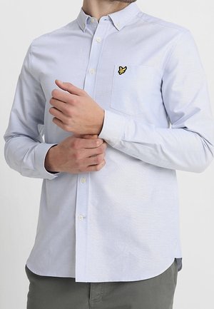 Un homme ajuste le poignet de sa chemise bleu clair à boutons avec un logo d'oiseau jaune sur la poche poitrine, portant un pantalon gris sur fond uni.
