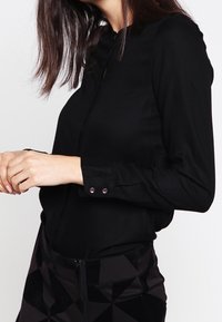 Black long-sleeve blouse with a buttoned collar and cuffs, made of smooth fabric. The wearer shows the blouse paired with a textured black skirt.