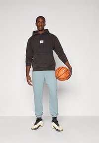 Man in black hoodie and light blue joggers holding an orange basketball against a plain white background.