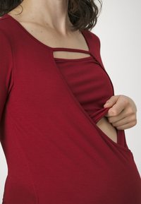 Raudona nėštuminė palaidinė su sluoksniuota struktūra, turinti apvalų iškirpimą ir reguliuojamą kryžminę dalį, užtikrinančią komfortą ir prieinamumą.