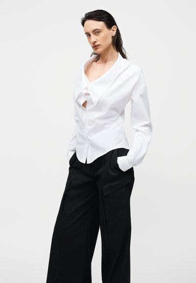 Γυναίκα με βρεγμένα μαλλιά που φοράει λευκό ασύμμετρο πουκάμισο με γιακά και μαύρο φαρδύ παντελόνι cargo, όρθια με τα χέρια στις τσέπες.