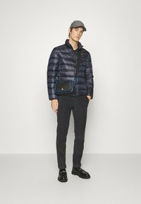 Giacca puffer blu navy con zip frontale, borsa a tracolla nera con dettagli blu, pantaloni scuri e scarpe nere lucide, indossati da un modello.