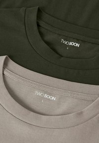 Två T-tröjor staplade; en olivgrön och en ljusgrå. Båda har runda, texturerade kragar och en etikett med texten "TWO SOON", storlek L.