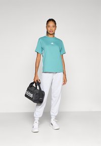 Türkise lühikeste varrukatega t-särk, millel on väike valge logo, koos heledate hallide dressipükstega ja musta duffelitaskuga, millel on valge bränding.