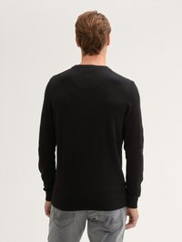 Hombre con cabello corto castaño que lleva un suéter negro de manga larga y jeans grises, de pie de espaldas frente a un fondo claro y liso.