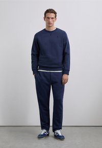 Sudadera azul marino con cuello redondo y puños acanalados, combinada con pantalones deportivos ajustados a juego. Acentuada con una capa interior gris claro y zapatillas deportivas.