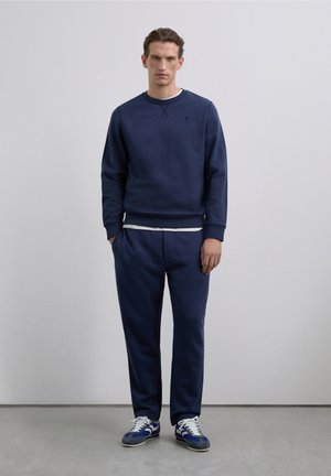 Sudadera azul marino con cuello redondo y puños acanalados, combinada con pantalones deportivos ajustados a juego. Acentuada con una capa interior gris claro y zapatillas deportivas.