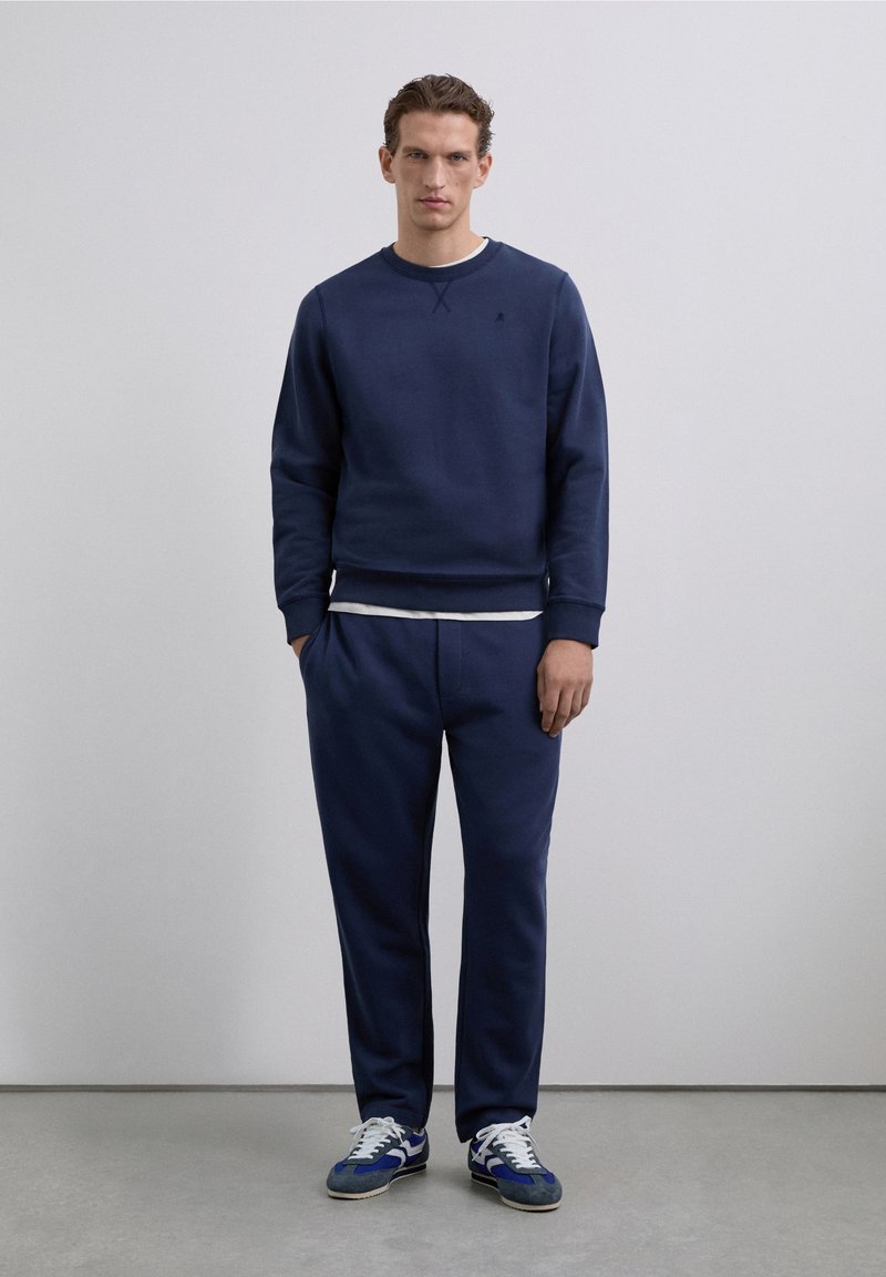 Sudadera azul marino con cuello redondo y puños acanalados, combinada con pantalones deportivos ajustados a juego. Acentuada con una capa interior gris claro y zapatillas deportivas.