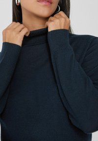 Suéter de cuello alto de un azul marino oscuro con textura acanalada, mangas largas y un cuello alto, sostenido por manos cerca del área del cuello.