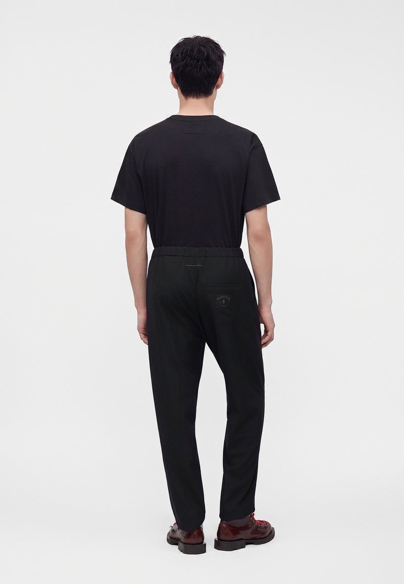 Homme debout, de dos, portant un t-shirt noir uni, un pantalon noir avec un patch sur la poche arrière et des chaussures marron foncé avec des lacets rouges.