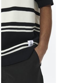 Le polo à manches courtes présente des rayures blanches et noires, un tissu texturé et un ourlet étiqueté. Porté avec un short sombre.